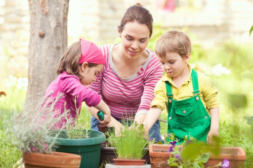 A woman with dark hair tied back, wearing a pink and white striped long-sleeve top, is outdoors in a garden with two children, one girl and one boy. The girl is dressed in a purple top and a pink headband, while the boy is wearing a yellow shirt and green dungarees. They are engaging in gardening activities with various pots and trays containing young plants and flowers, including lavender and other flowering species. The garden setting features a large tree trunk in the background, along with a hedge and a brick wall, suggesting a landscaped backyard in Southfields, London. The scene is illuminated by soft natural sunlight, indicating a pleasant, mild day suitable for outdoor gardening work. The image reflects a family-friendly gardening activity, emphasizing nurturing plants and enjoying outdoor landscaping, which aligns with professional gardening and lawn care services offered by Lawn Mowing Southfields in the local area, including Southfields postcodes and surrounding neighborhoods.
